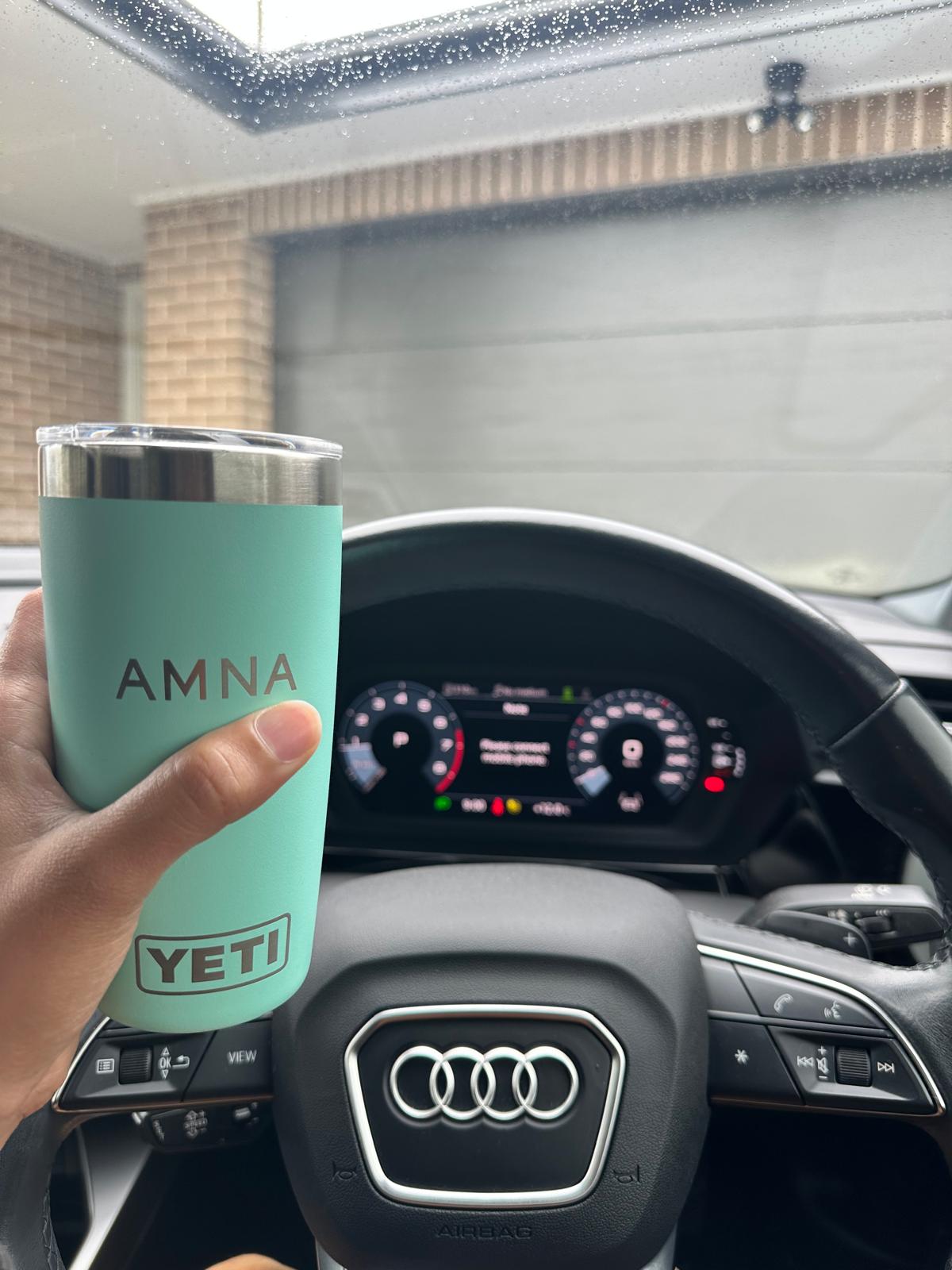 A mint-green travel mug engraved with Amna’s name, held in the car on a rainy day